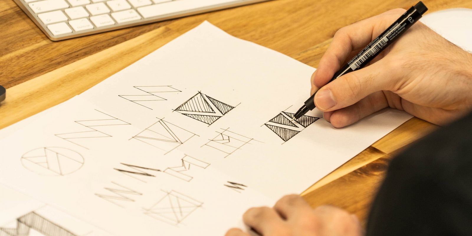 Designer's hand sketching logo ideas with a pen on paper next to a keyboard
