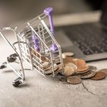 Miniature shopping cart tipped over with coins beside an open laptop