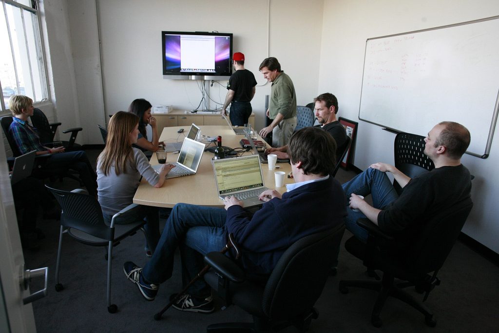 Small team seated around a meeting table with laptops and a wall screen