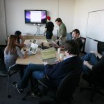 Small team seated around a meeting table with laptops and a wall screen