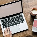 Top view of hands working on a laptop beside a smartphone, notebook and coffee on a wooden desk